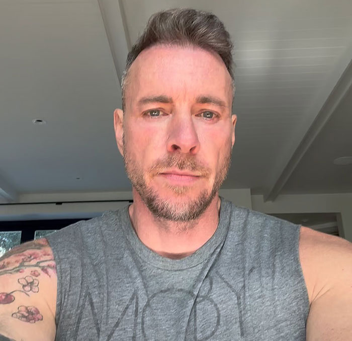 Dax Shepard in a sleeveless shirt, showing tattoos, appearing relaxed in a well-lit indoor setting.